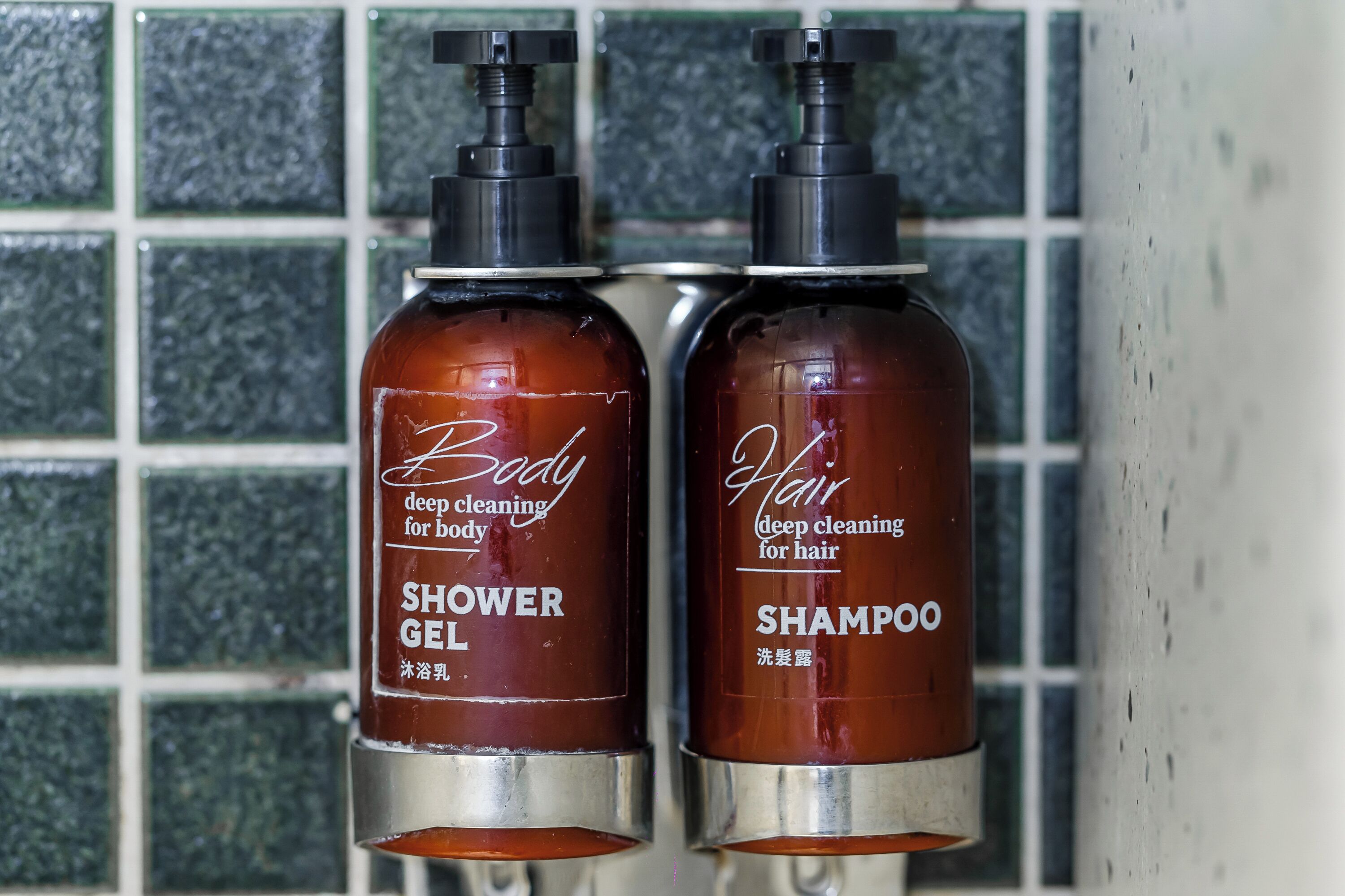 Two bottles of body and hair care products, labeled 'Shower Gel' and 'Shampoo', against a tiled wall.