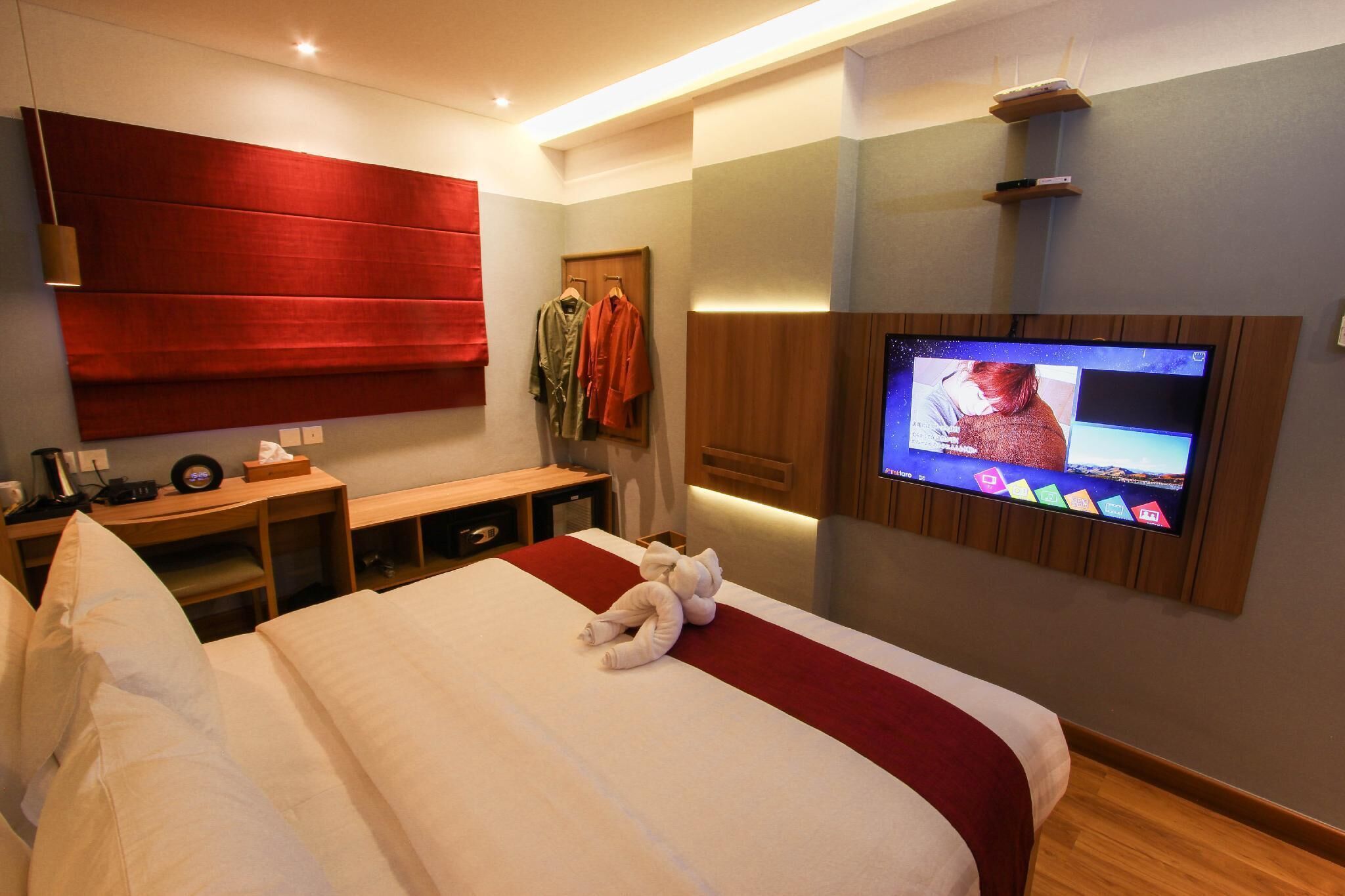 A hotel room with a large bed, a flat-screen TV mounted on the wall, a wooden desk, and a red headboard.