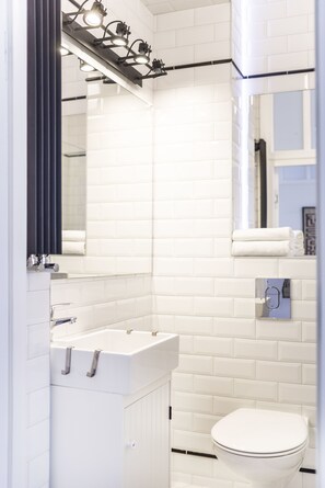 Une salle de bain moderne avec des carreaux de céramique blancs rectangulaires, un lavabo blanc avec un miroir au-dessus, et des toilettes blanches.