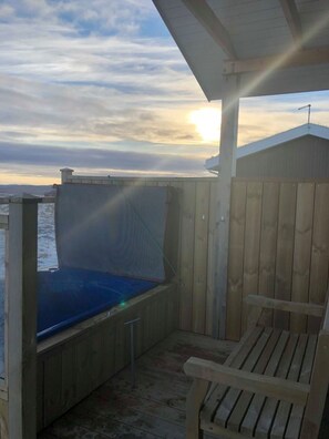 Une terrasse en bois avec un jacuzzi et une chaise, donnant sur un paysage enneigé au coucher du soleil.