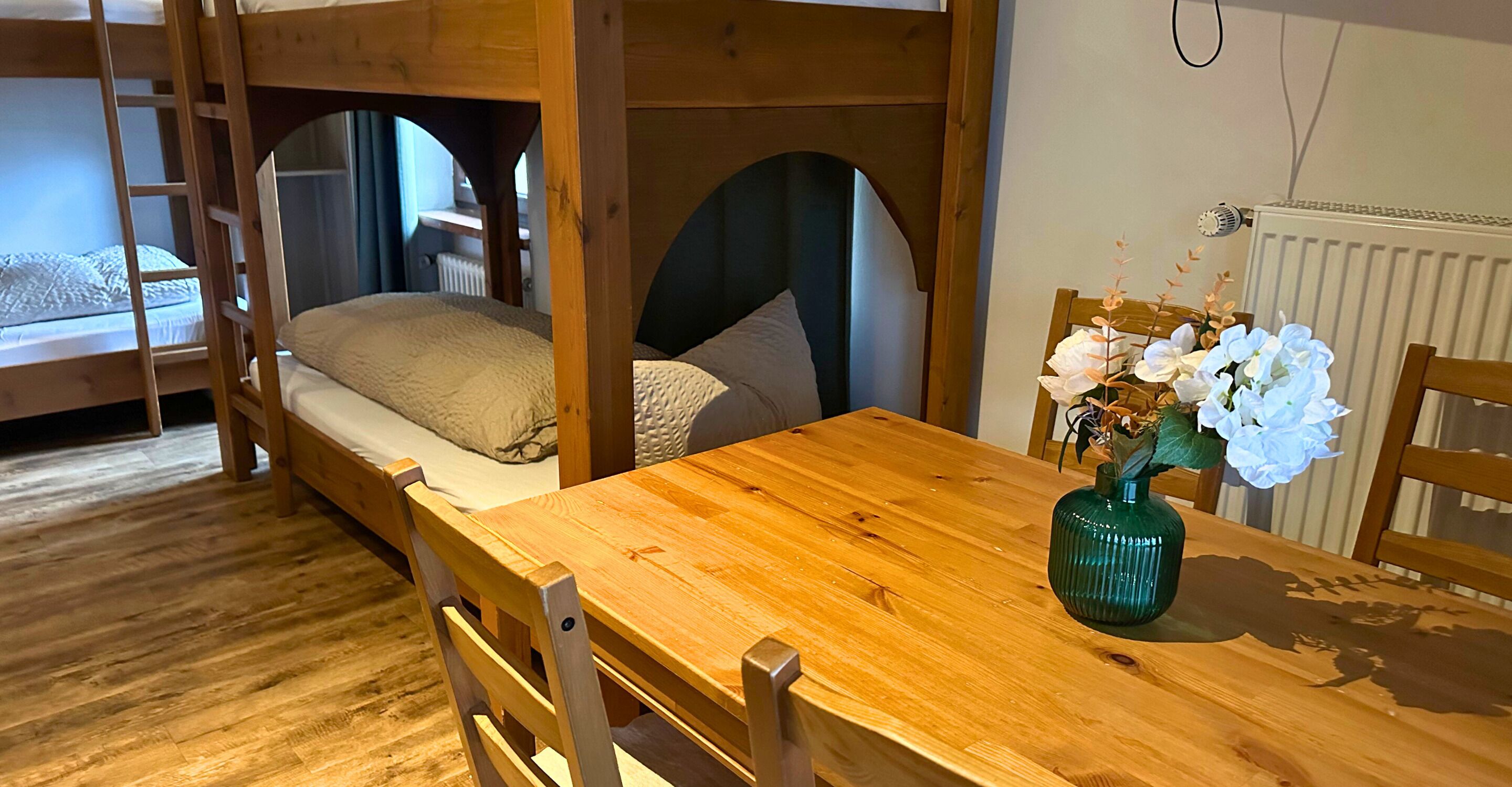 Un lit superposé avec une échelle, une table en bois ornée d’un vase de fleurs et une chaise.