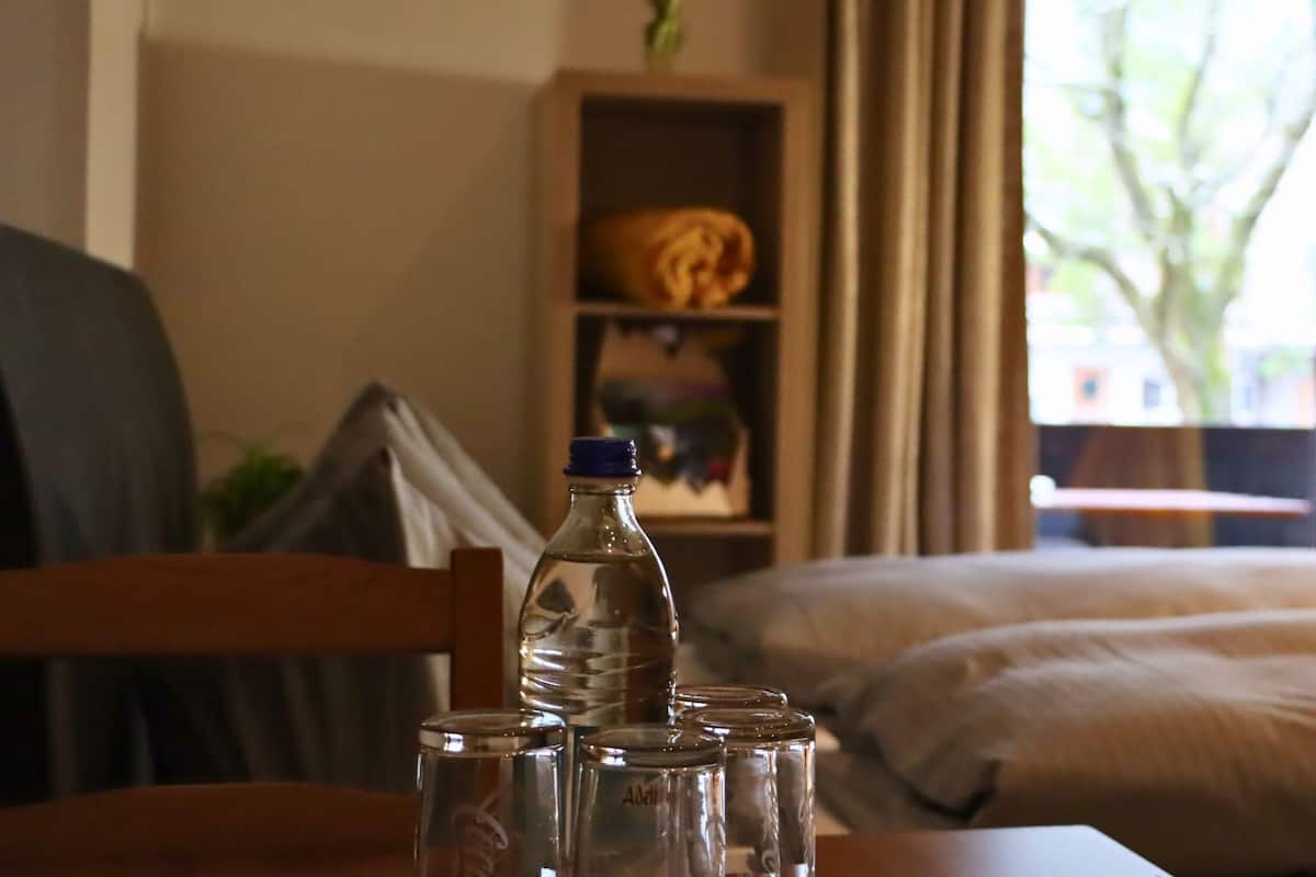 A hotel room with a bed, a wooden chair, a table with a water bottle and glasses, and a window with a view of a tree and buildings.
