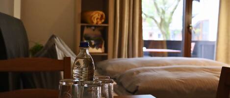 Une chambre d’hôtel avec un lit, une chaise en bois, une table avec une bouteille d’eau et des verres, et une fenêtre donnant sur un arbre et des immeubles.