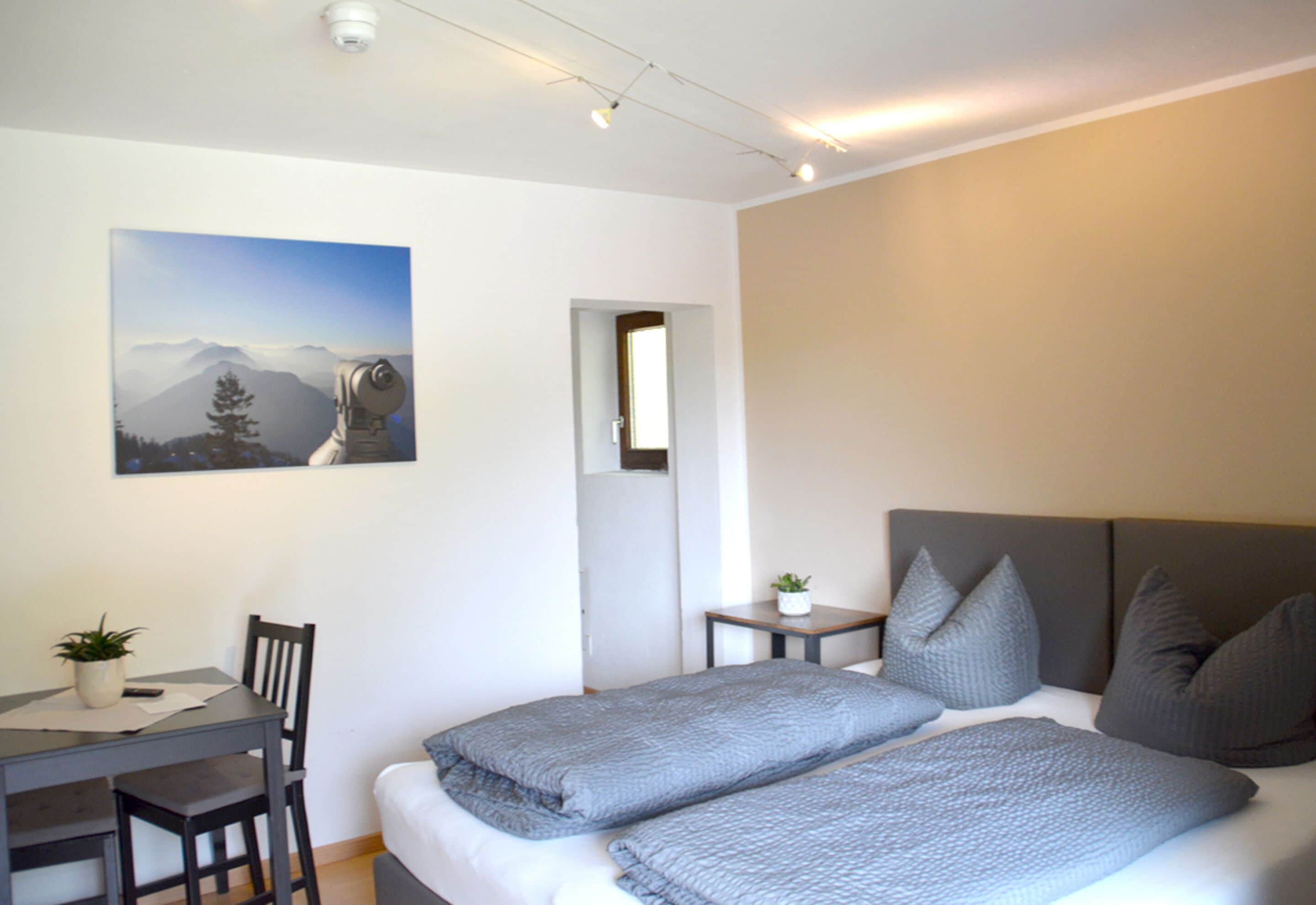 A bedroom with a bed, a desk, a chair, a small table, and a potted plant.