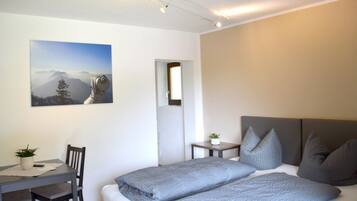 A bedroom with a bed, a desk, a chair, a small table, and a potted plant.