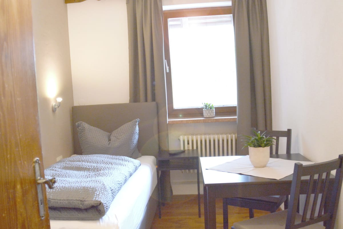 A bedroom with a bed, a bedside table, a window with curtains, a radiator, and a small potted plant on the windowsill.