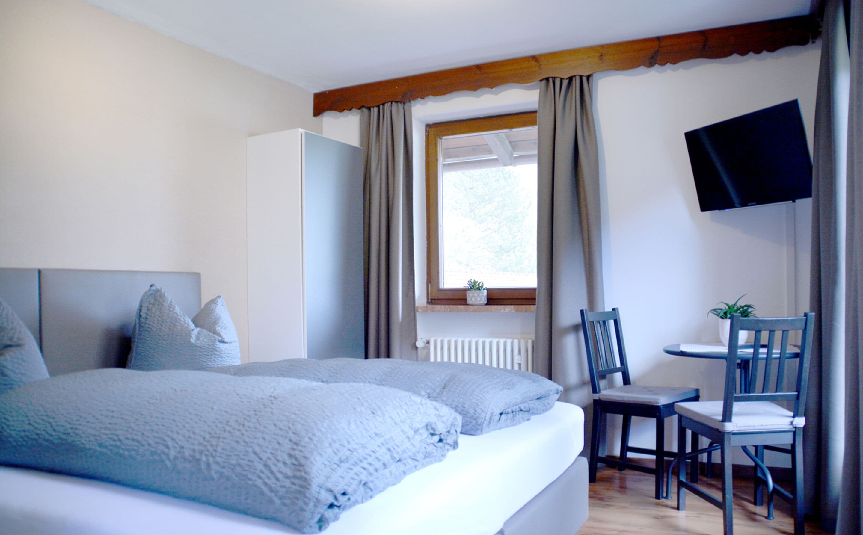 A bedroom with a bed, a desk with two chairs, a TV, and a window with curtains.