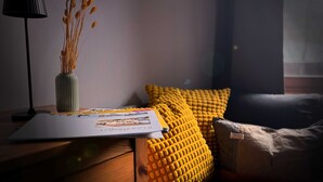 A room with a wooden nightstand, a lamp, a vase with dried plants, a magazine, a yellow textured pillow, and a dark sofa.