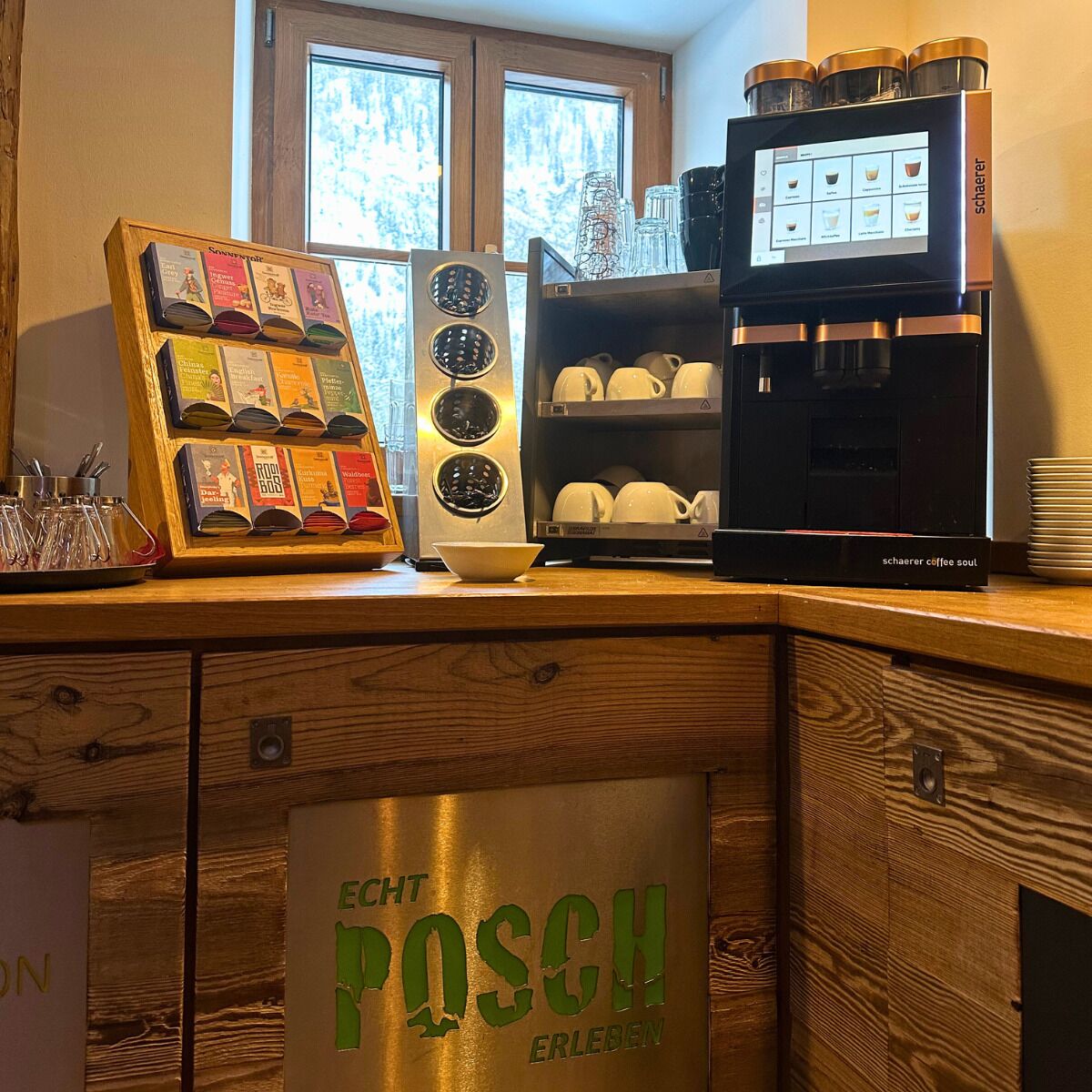 En kaffestation med en kaffemaskin, ett utbud av kaffekapslar och en skylt med texten ”Echt Posch Erleben”.