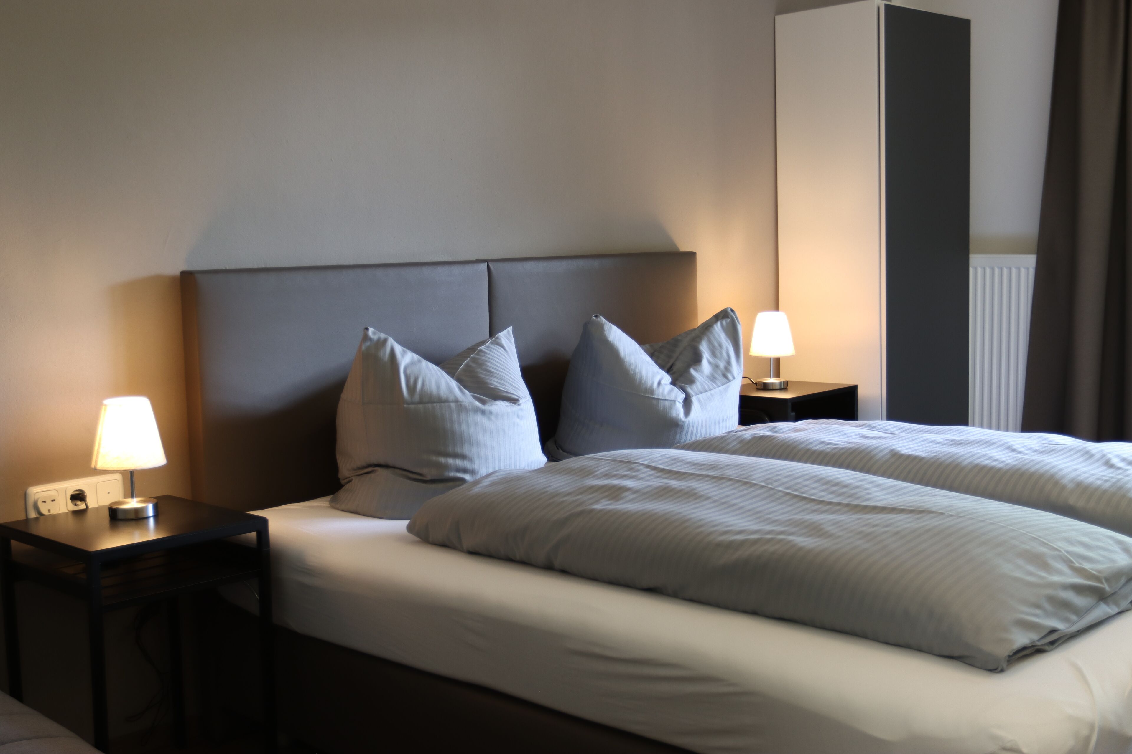 Una cama bien hecha con dos almohadas, una mesita de noche con lámpara y un armario al fondo.