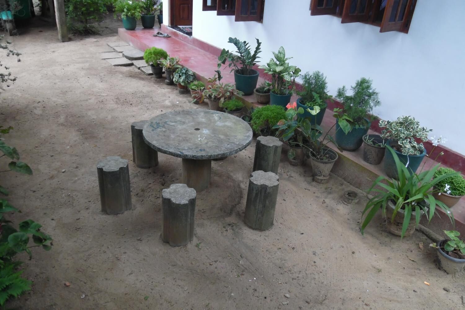 Un patio con una mesa redonda rodeada de troncos de madera, plantas en macetas y un edificio blanco con detalles rojos.
