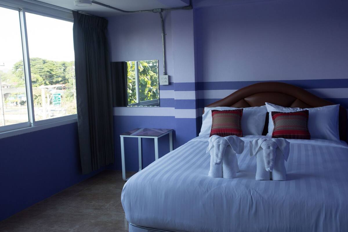 Ein Hotelzimmer mit einem großen Bett, einem kleinen Tisch, einem Fenster mit Vorhängen und Blick auf Grünfläche.