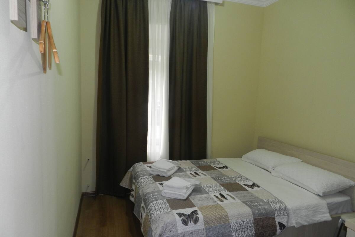 A single bed with a patterned bedspread, a white pillow, and a folded towel on the bed. There is a window with dark curtains partially drawn, and a wooden floor.