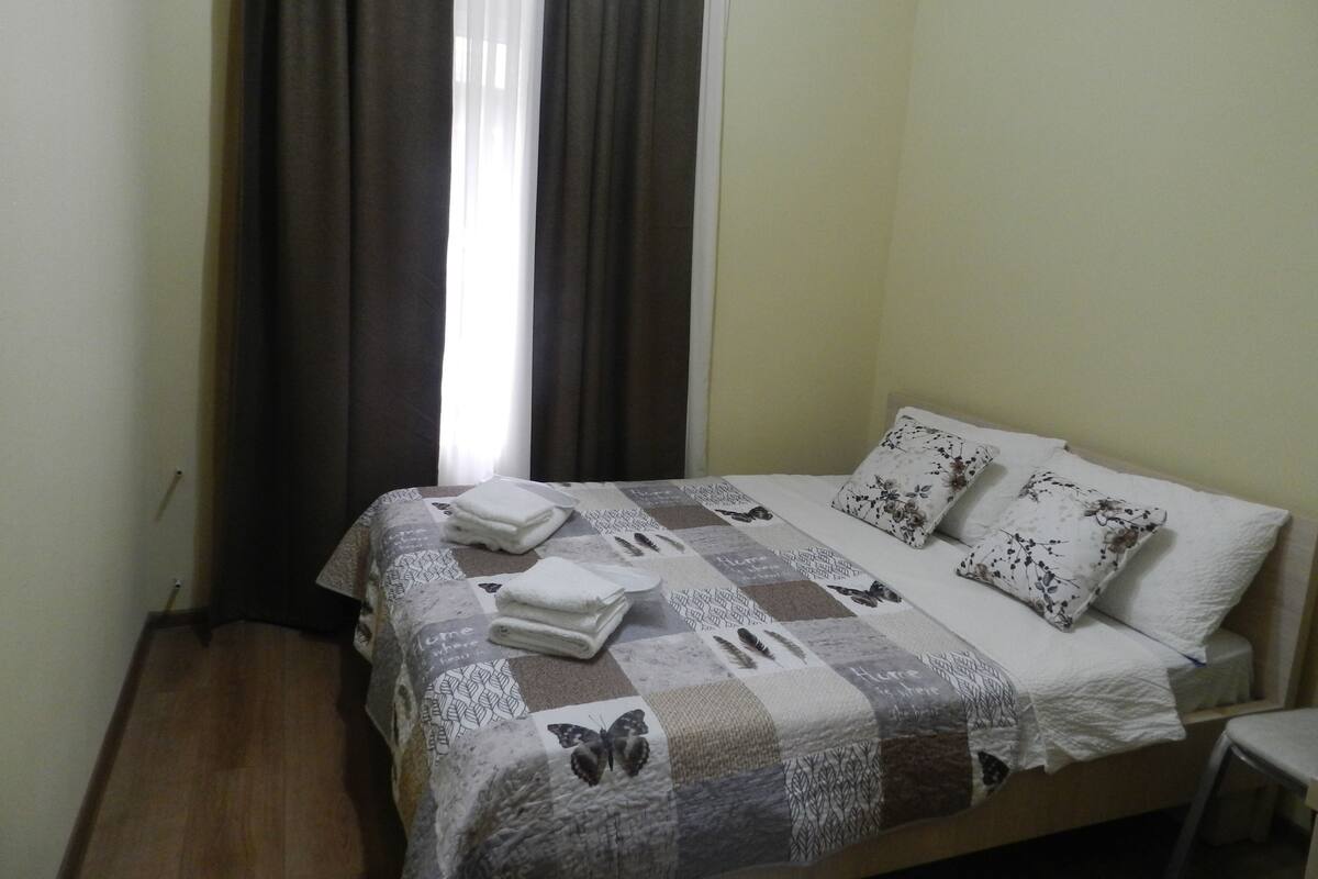 A neatly made bed with a patterned bedspread, two pillows with floral designs, and a folded towel on the bed.