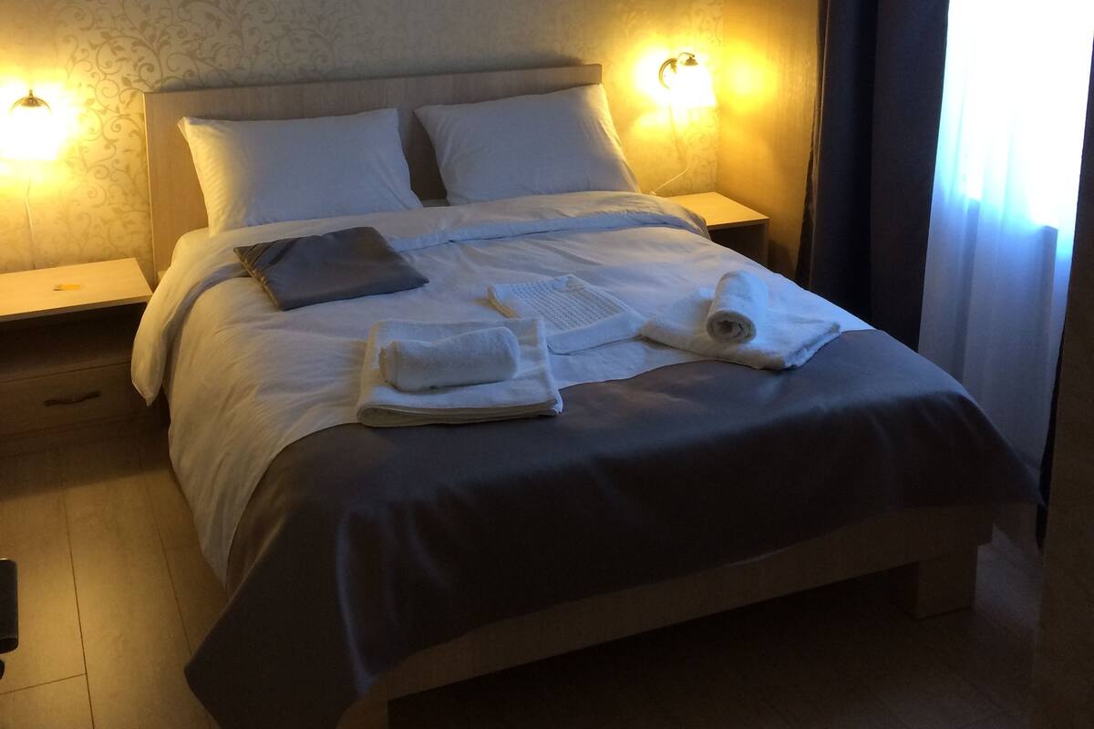 A neatly made bed with white linens and pillows, two bedside lamps, and a window with curtains.