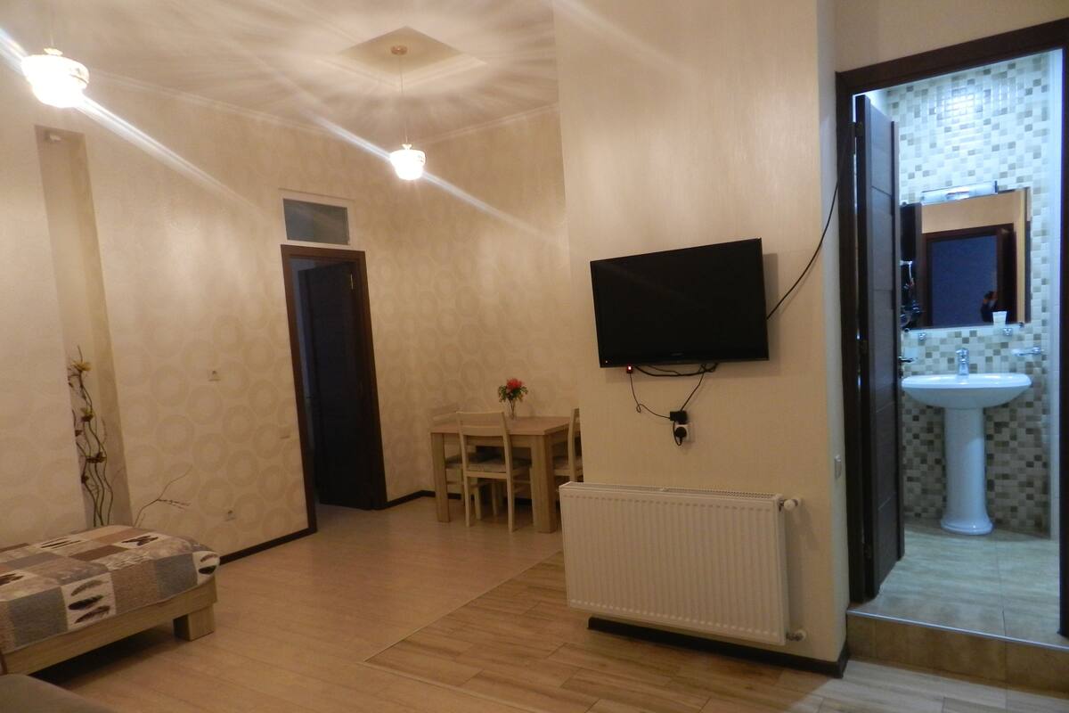 A living room with a TV mounted on the wall, a dining table with chairs, a radiator, and a sofa.