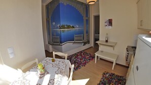 A room with a view of a blue sky and water through a window, a dining table with chairs, a bench, a kitchen area with cabinets, and a hanging light fixture. - Tango - Apartments on the Beach (Corfu)