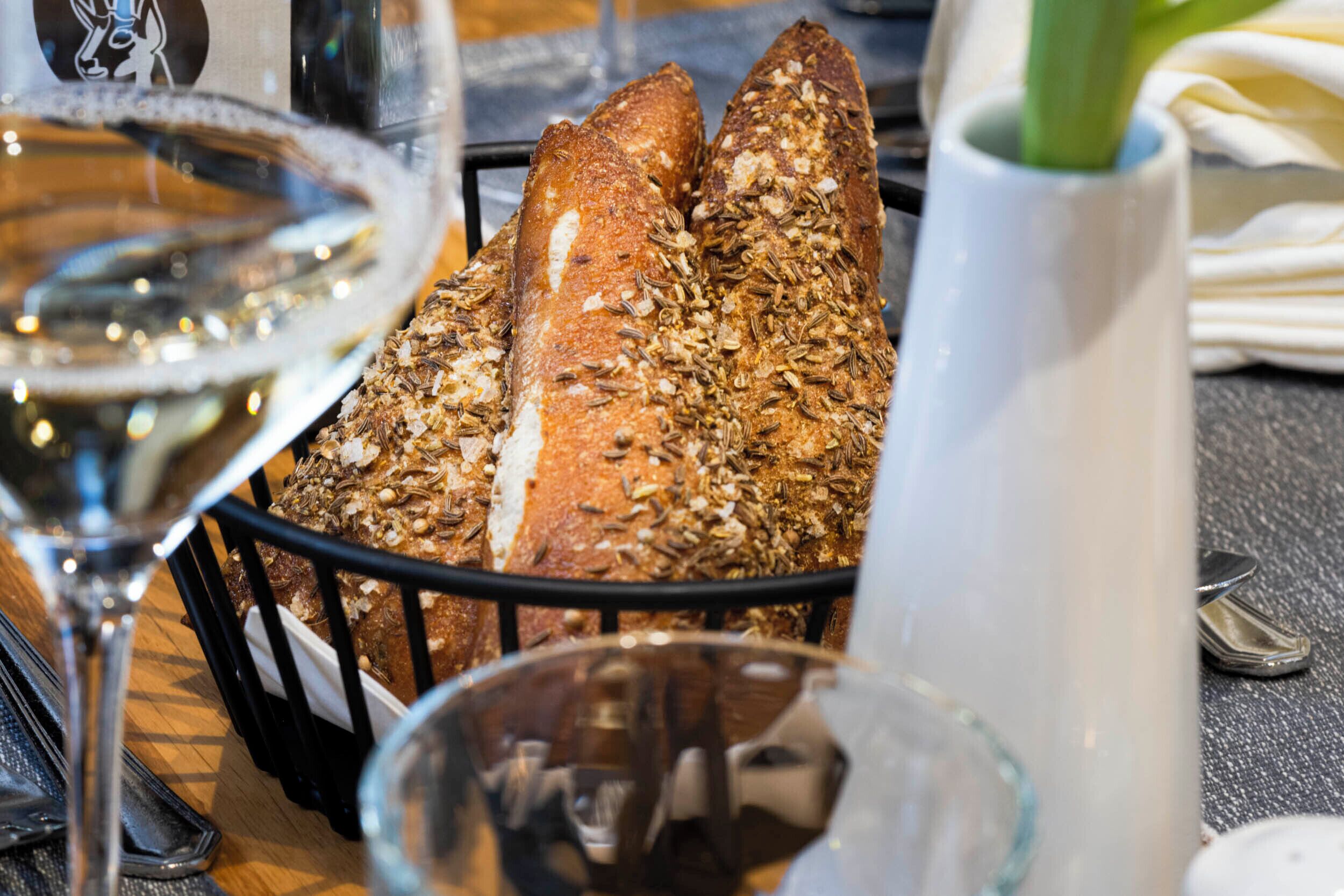 Un cesto di baguette con semi su un tavolo, un bicchiere di vino bianco e un vaso con una pianta verde.