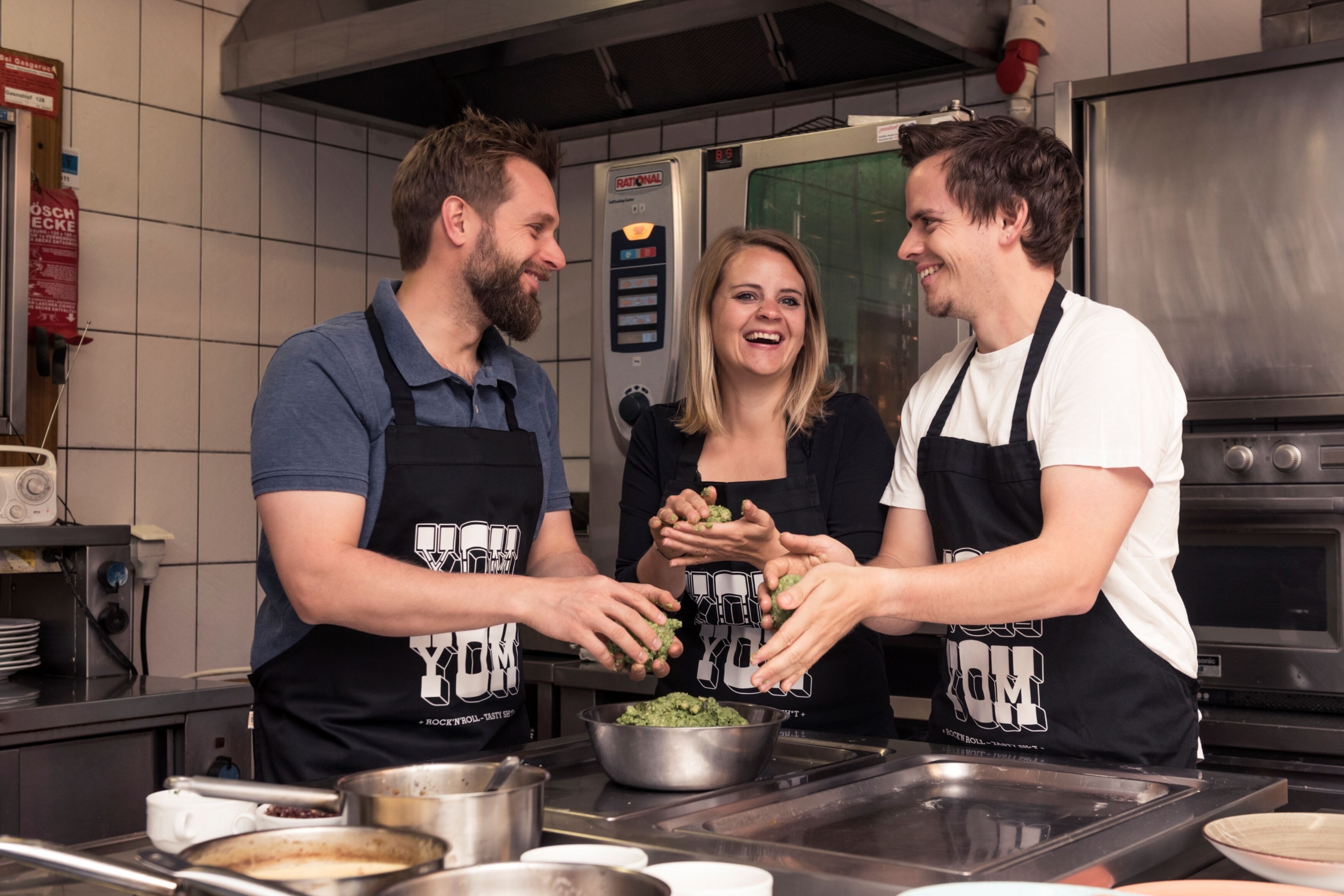 Tres personas en una cocina preparando alimentos, con electrodomésticos de acero inoxidable y un salpicadero de azulejos.