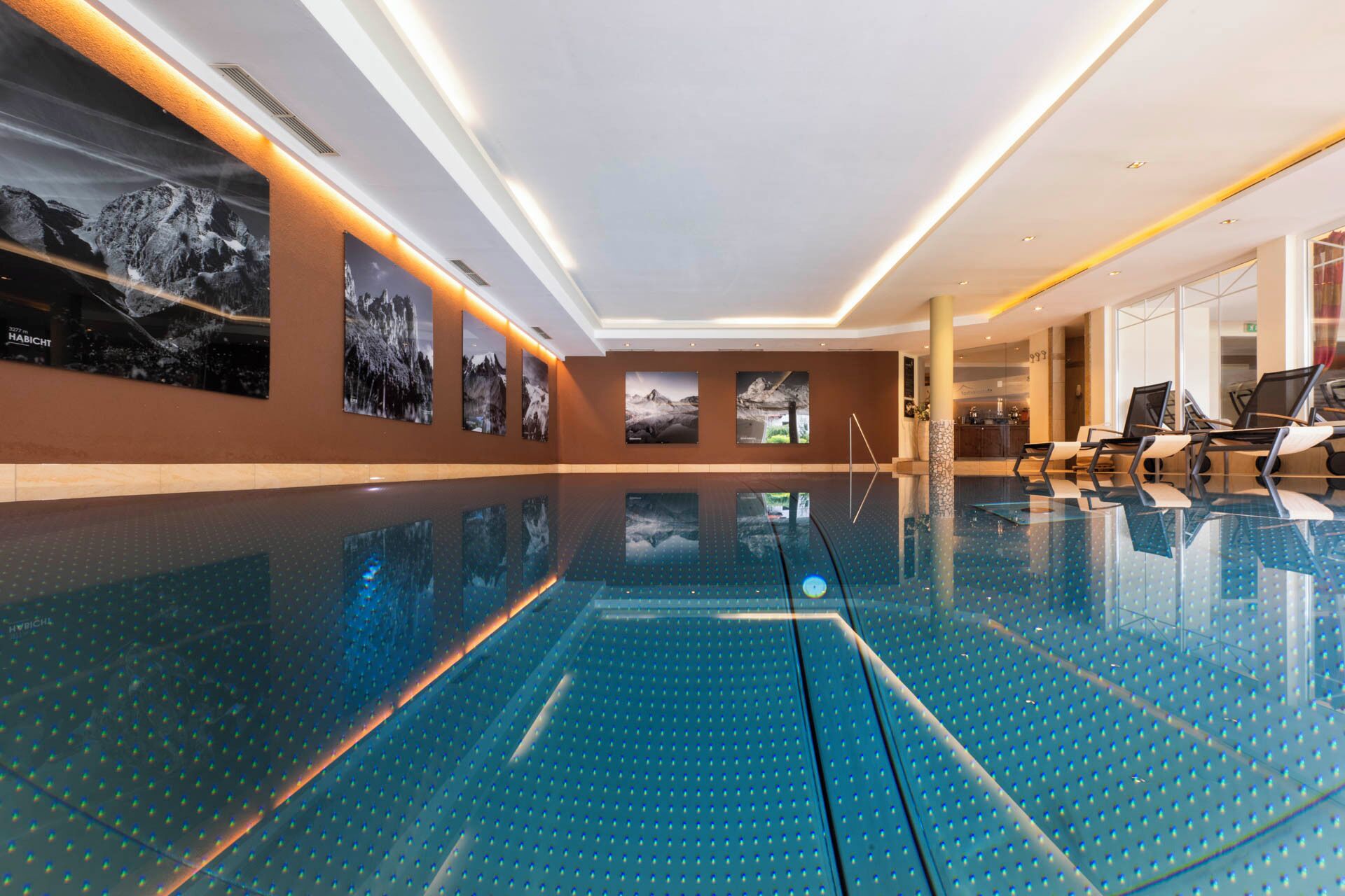 A modern indoor swimming pool with a clear blue floor, surrounded by walls with framed photographs.