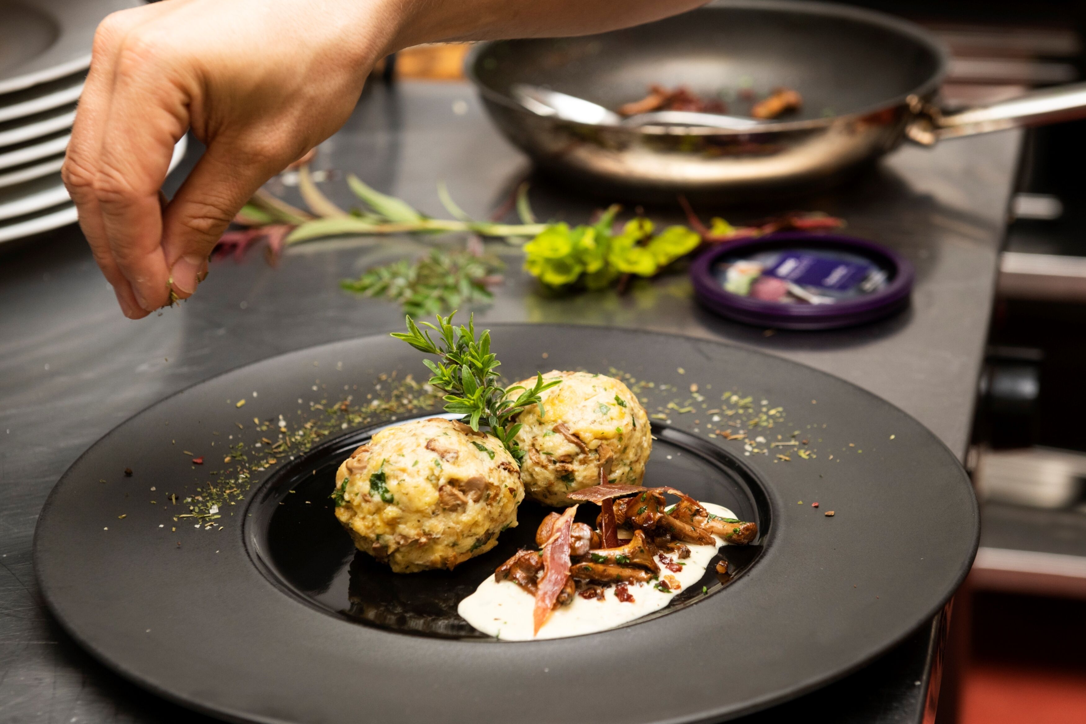 La main d’un chef dépose une branche d’herbes aromatiques sur une assiette contenant deux raviolis et une sauce.