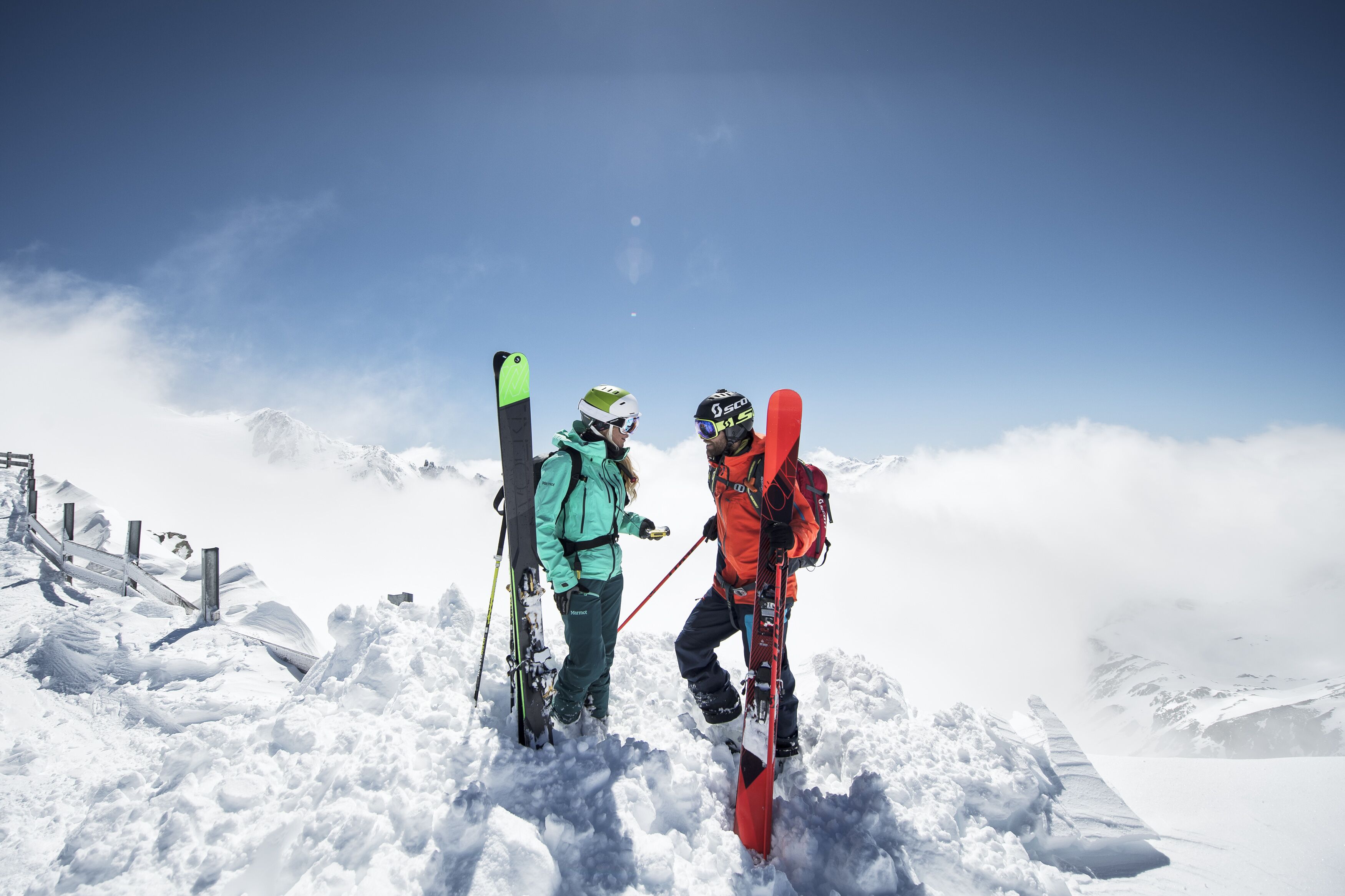 Zwei Skifahrer auf einem verschneiten Berg unter blauem Himmel.
