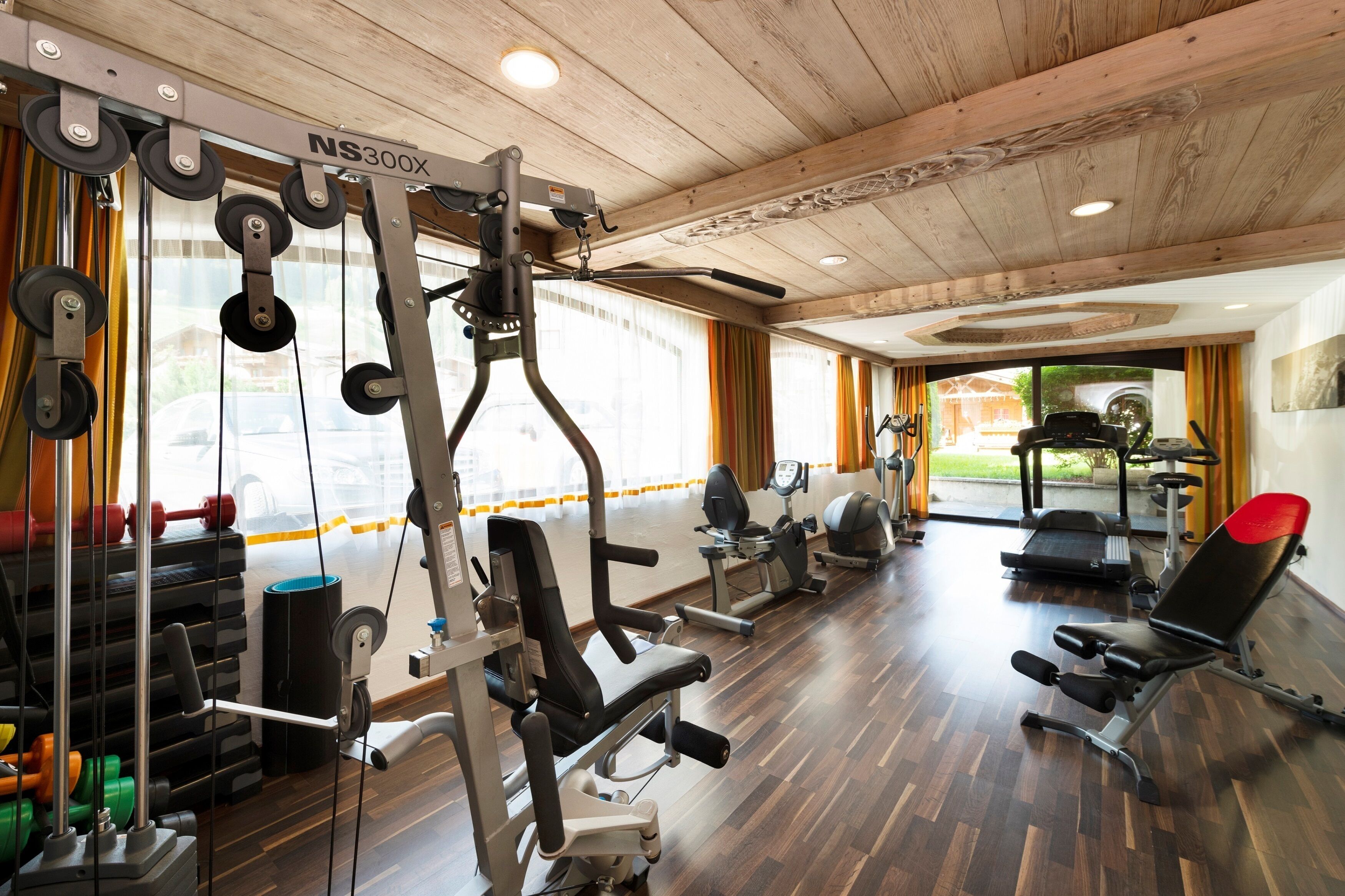 Et fitnessrum med forskellig træningsudstyr, trægulve og store vinduer med gardiner.