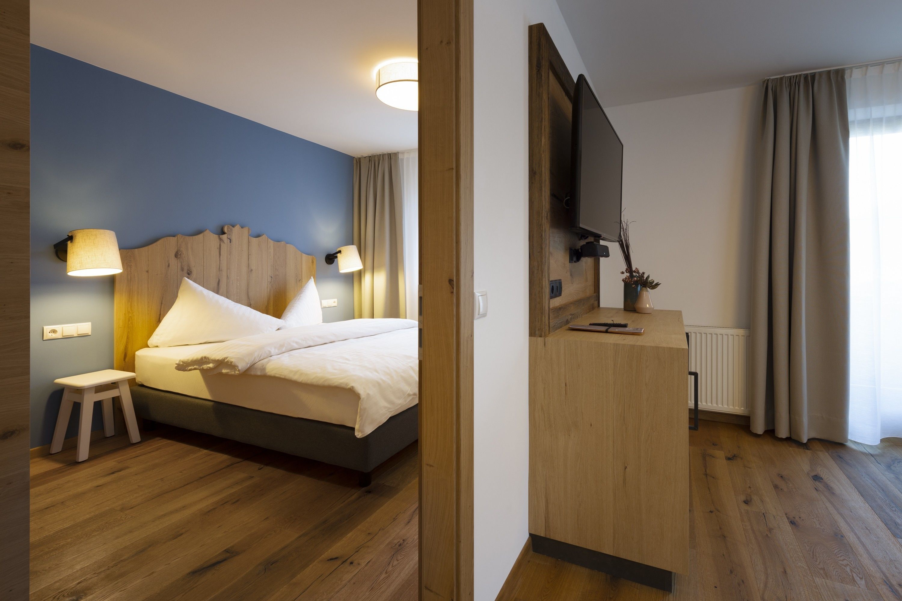 A modern hotel room with a wooden floor, a bed with white linens, a wooden headboard, a small wooden stool, a bedside table with a lamp, a TV mounted on the wall, and a window with curtains.