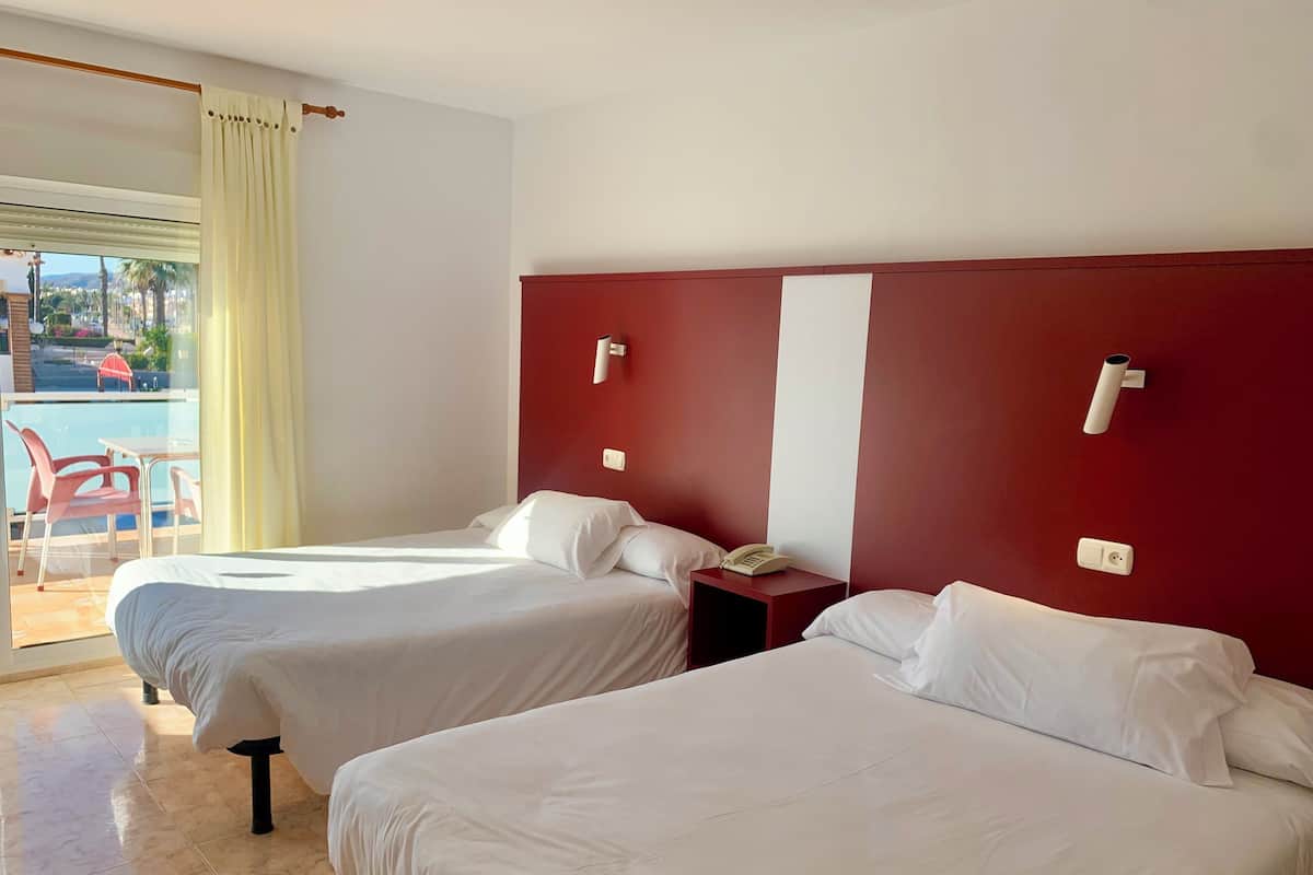 A hotel room with two beds, a red accent wall, a small bedside table with a phone, and a view of a pool and outdoor seating.