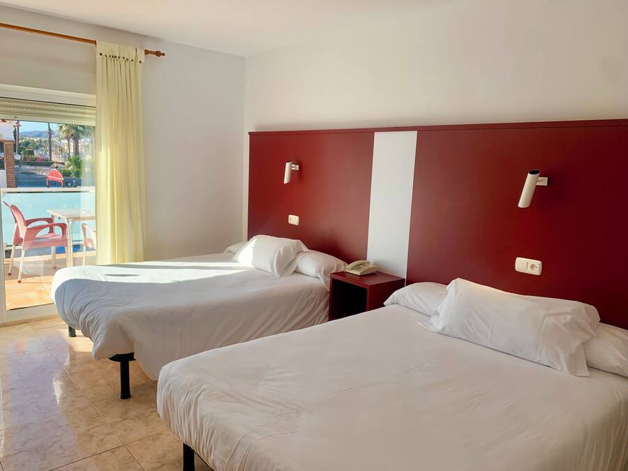 A hotel room with two beds, a red accent wall, a small bedside table with a phone, and a view of a pool and outdoor seating.