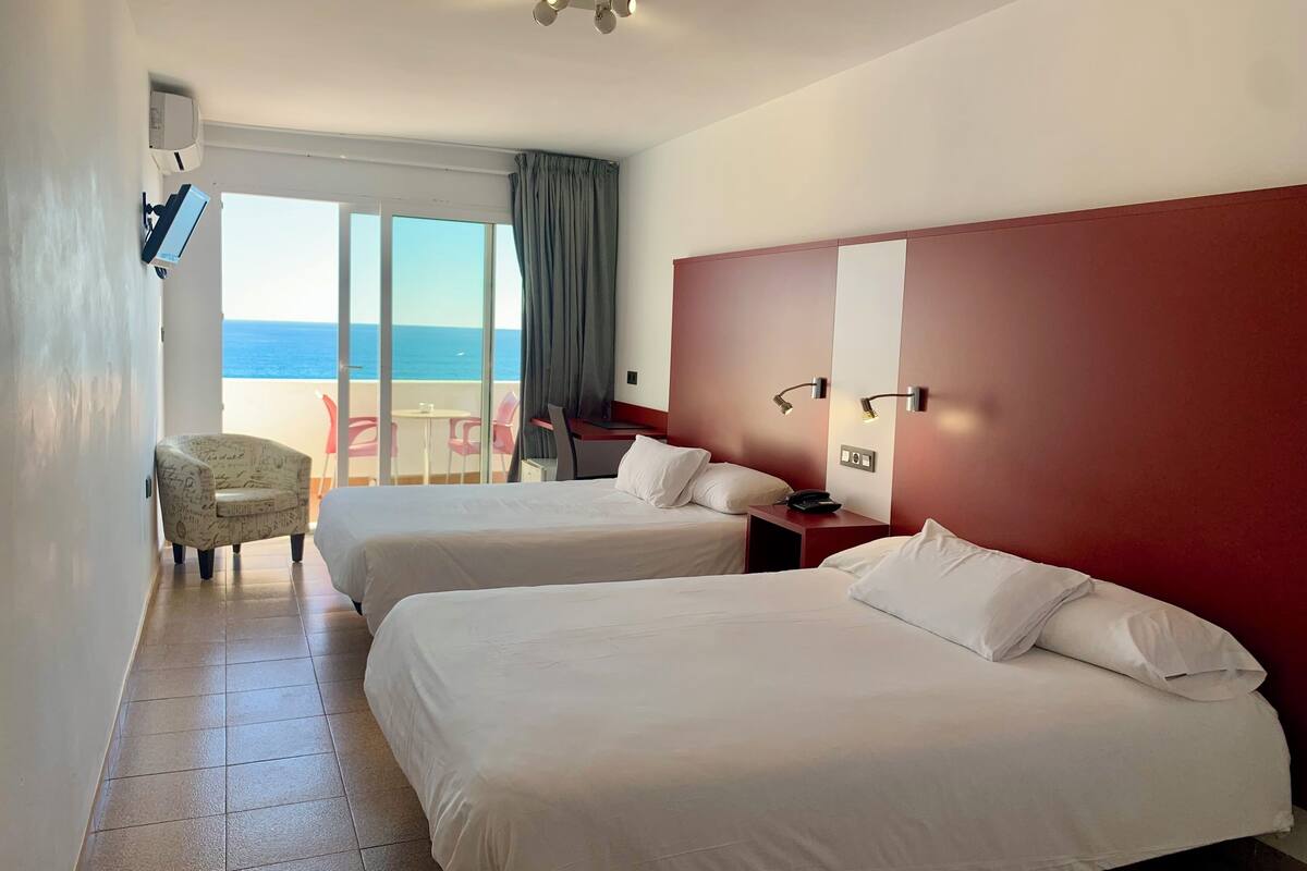 A hotel room with two beds, a desk, a chair, and a view of the sea.