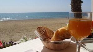 Een tafel met een broodmand en een glas sinaasappelsap, met uitzicht op een strand.