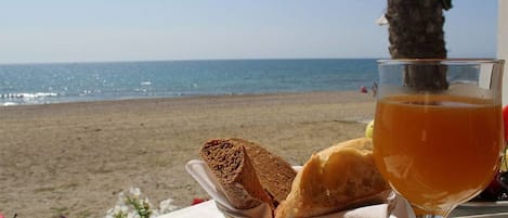 A table with a basket of bread and a glass of orange juice, overlooking a beach.