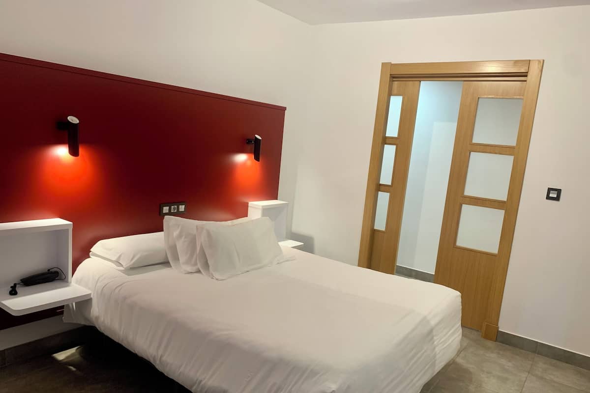 A neatly made bed with white linens in a modern room with a red accent wall and two bedside lamps.
