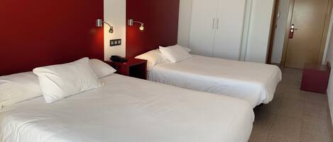 Quarto de hotel com duas camas, uma parede de destaque vermelha, um guarda-roupa e uma mesa de cabeceira com telefone.