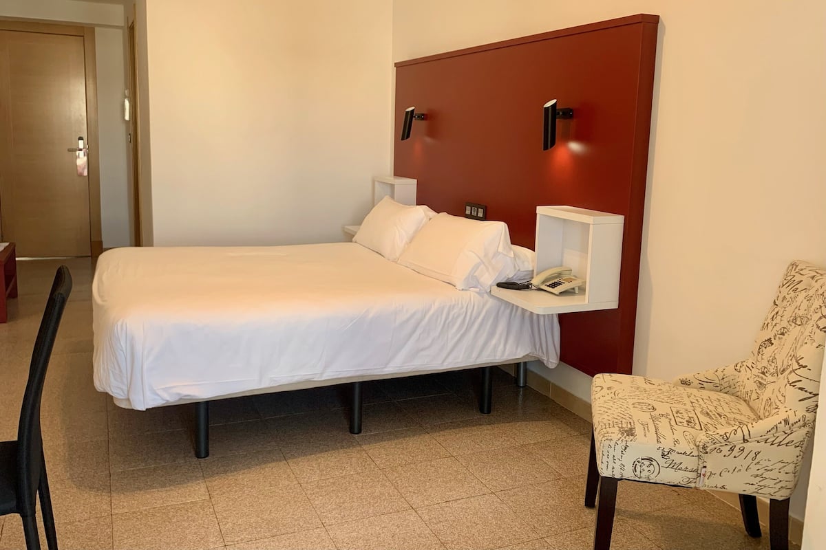 A hotel room with a bed, a chair, a nightstand with a phone, and a red accent wall.