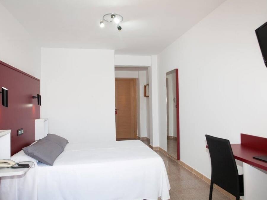 Habitación de hotel con cama, mesita de noche, lámpara, escritorio, silla y televisor montado en la pared.