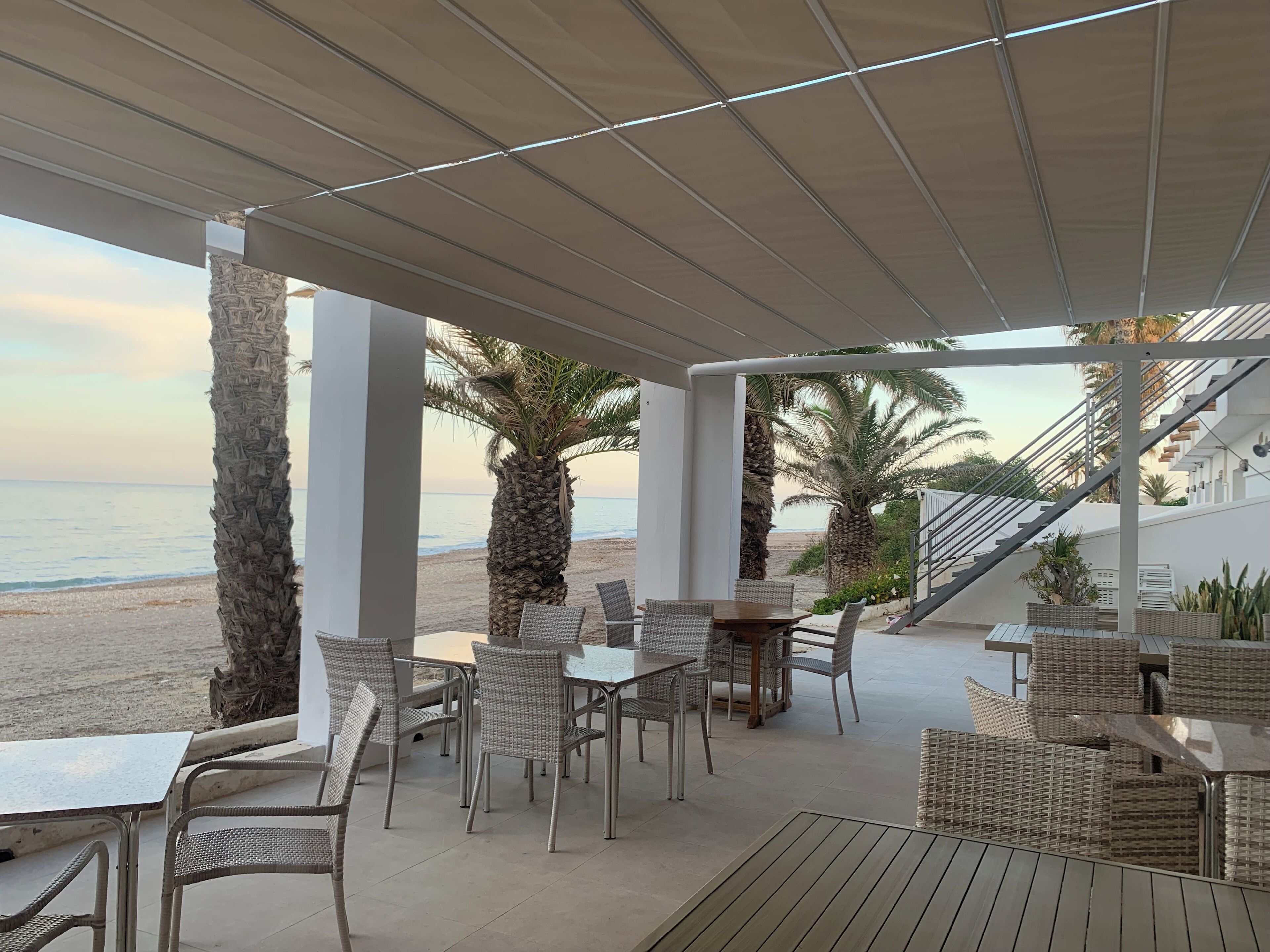 Eine Terrass mit Tischen und Stühlen mit Blick auf einen Strand, gesäumt von Palmen, und einer Treppe, die zu einem Gebäude führt.