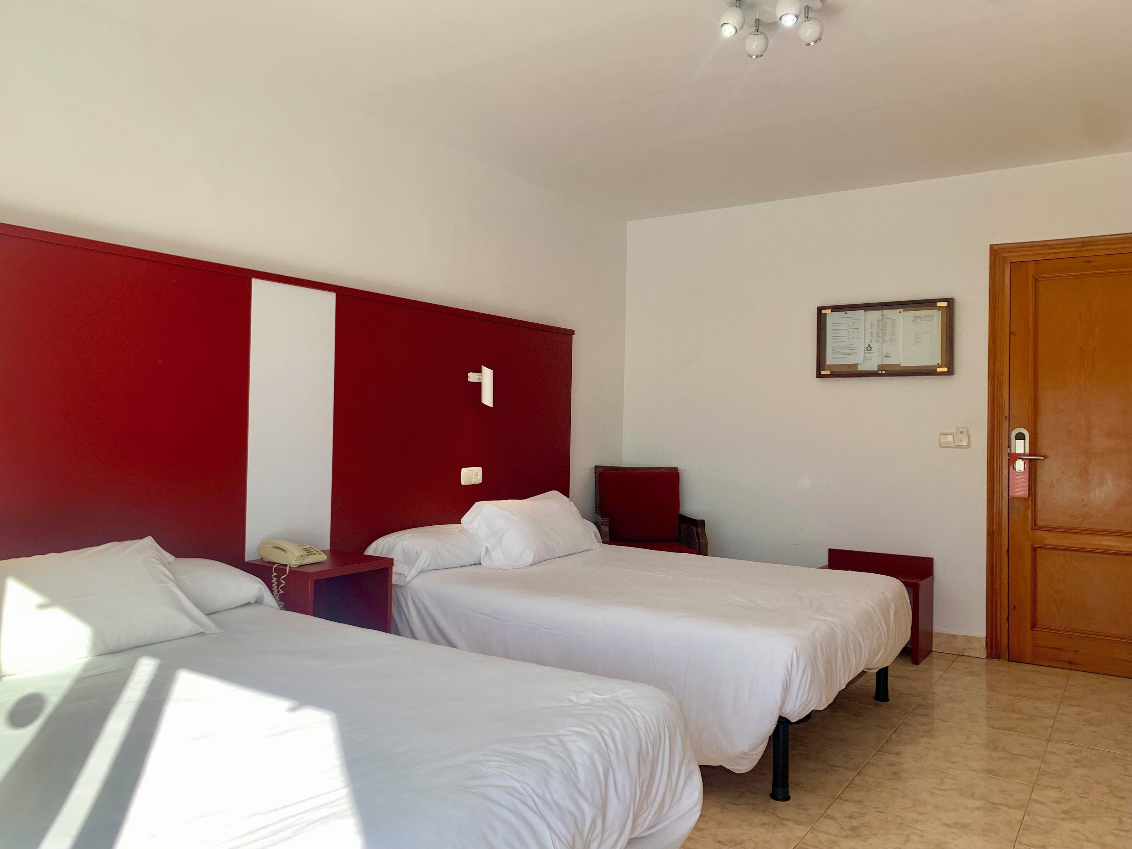 Una habitación de hotel con dos camas individuales, una pared de acento roja, una puerta de madera y un aviso enmarcado en la pared.