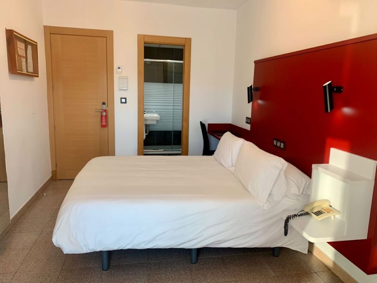 Una habitación de hotel con una cama grande, un armario rojo, un teléfono en la mesita de noche y una puerta al baño.