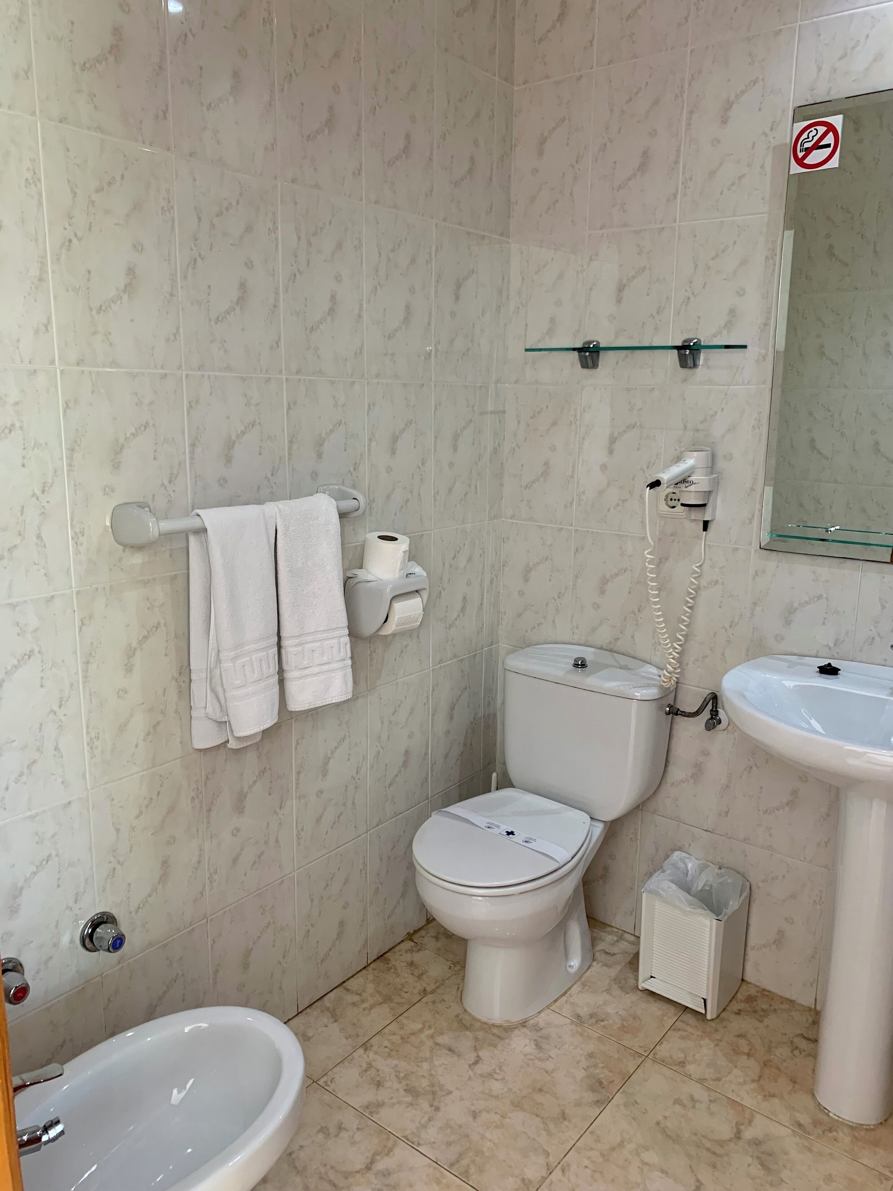 A bathroom with a toilet, bidet, sink, and a glass shelf above the toilet.