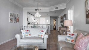 A spacious living room with a white sofa, a glass coffee table, and a beige sofa with decorative pillows. A chandelier hangs from the ceiling, and artwork is displayed on the walls. - Beach Club Bristol 202 2 Bedroom Condo by RedAwning (Gulf Shores)