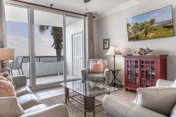 A living room with a glass table, two sofas, a patterned armchair, a red cabinet, and a painting of a beach scene.
