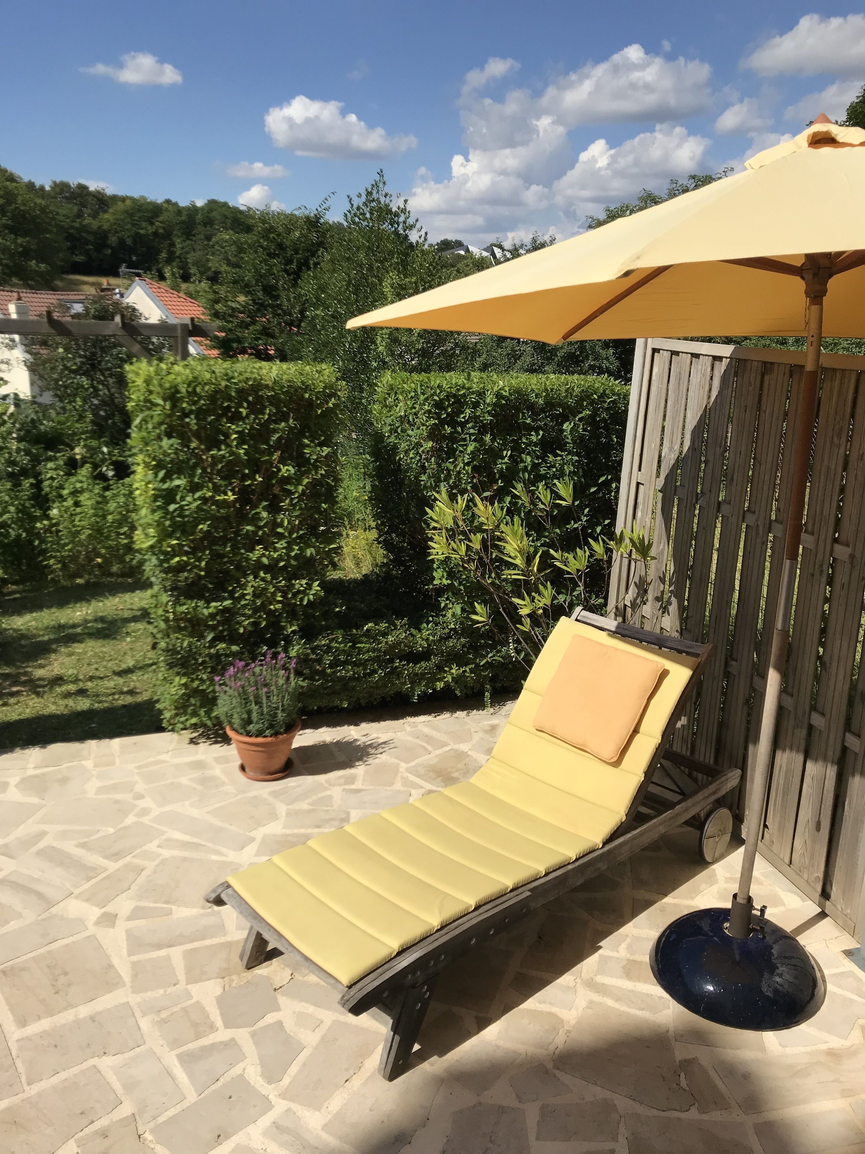 Eine Sonnenliege mit gelber Auflage und blauem Sonnenschirm, platziert auf einer Steinterrasse mit Holzlattenzaun und Grünpflanzen im Hintergrund.