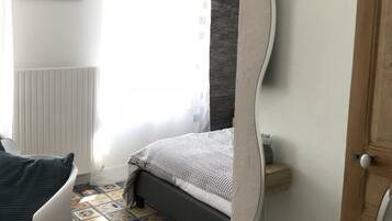 A bedroom with a bed, a chair, a radiator, a window with curtains, and a wall-mounted mirror.