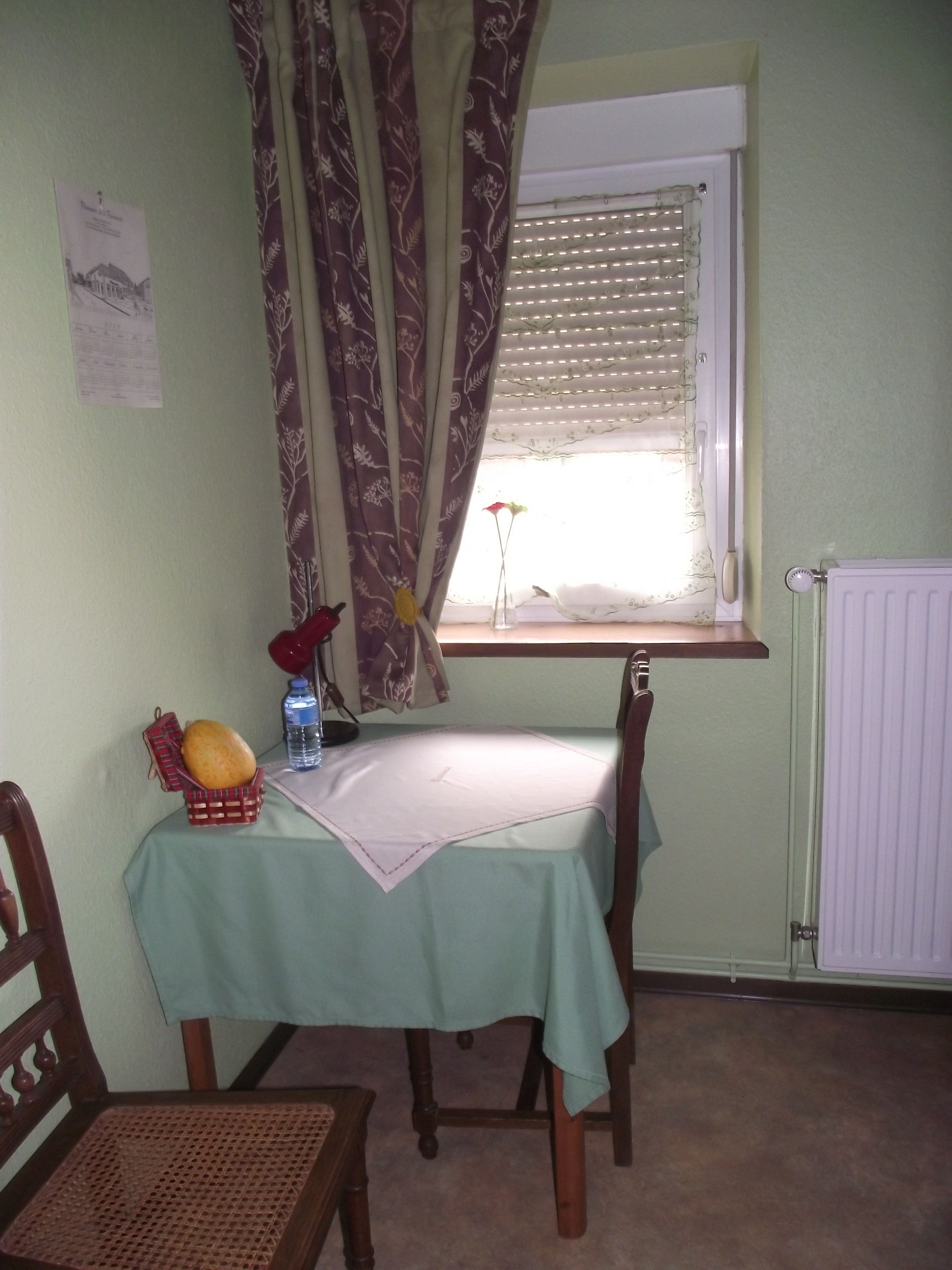 A small dining area with a table set for two, a chair, a window with blinds, a radiator, and a curtain.