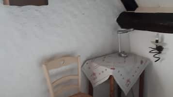 Ett litet rum med en trästol, ett litet bord med en duk, en väggklocka och en säng med ett mönstrat överkast.