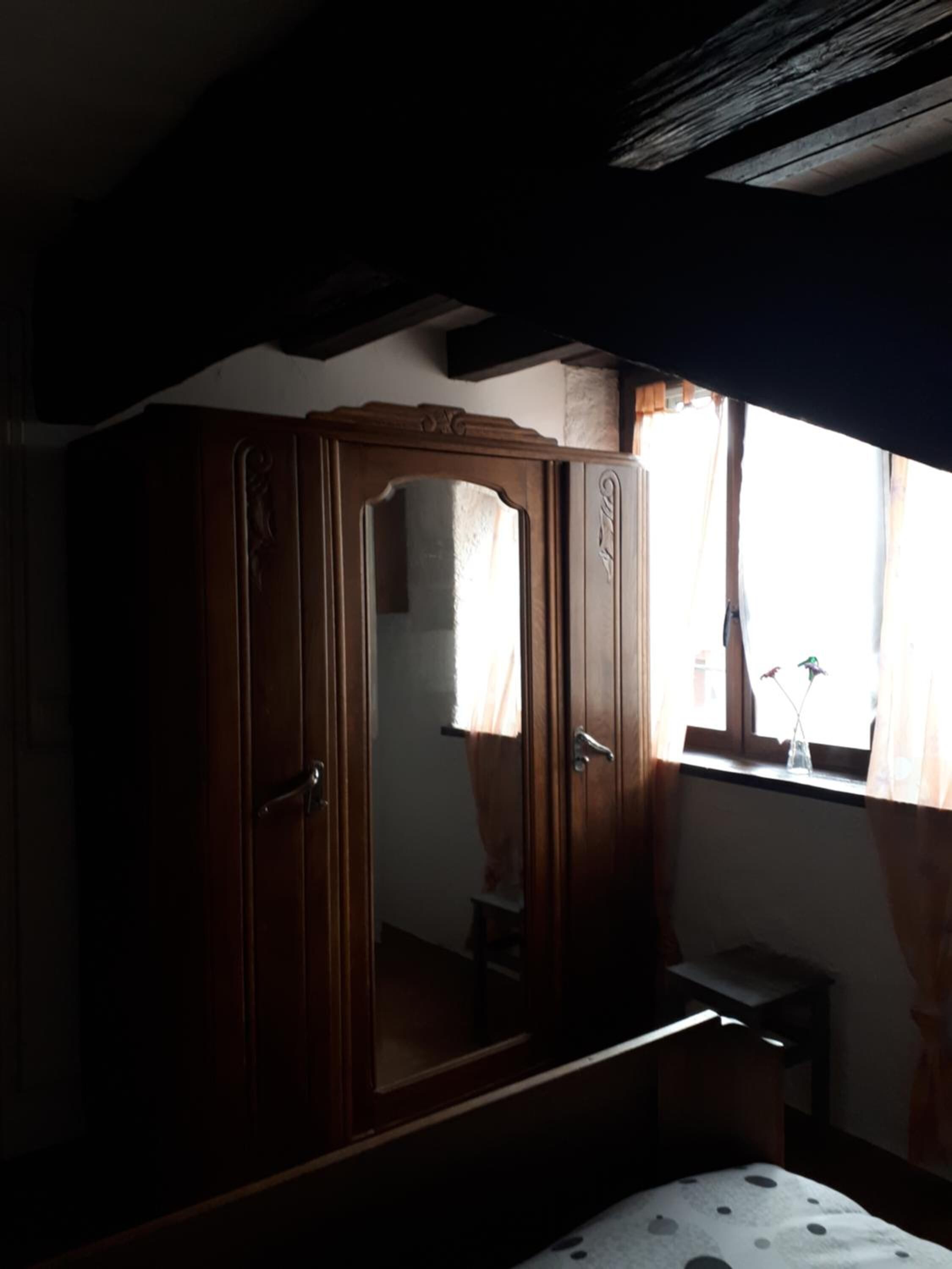 A bedroom with a wooden wardrobe, a small bedside table, and a window with curtains.