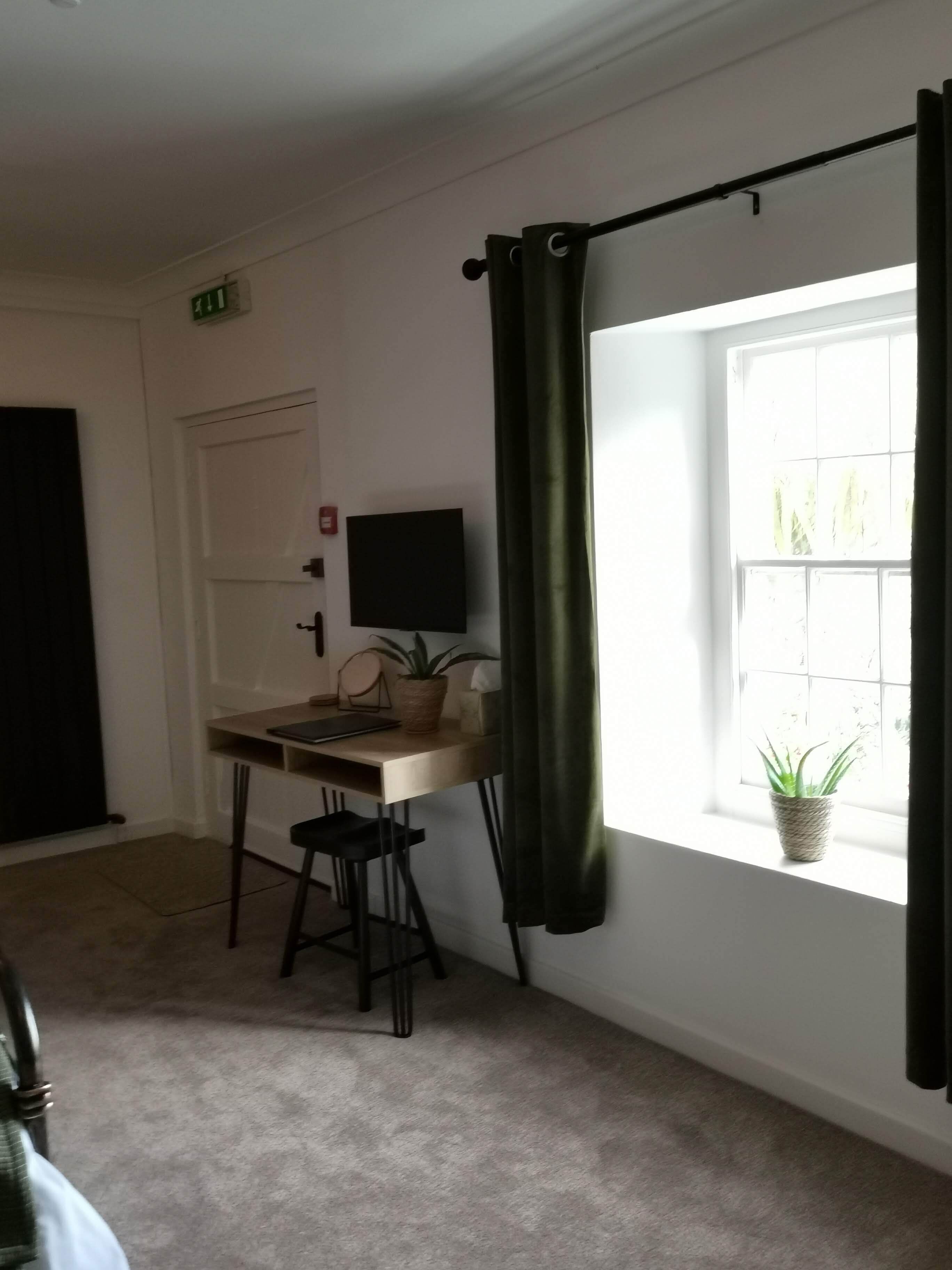 Ein Zimmer mit Schreibtisch, Stuhl, Fernseher, einer Zimmerpflanze und einem fensterbepflanzten Fenster mit Vorhängen.