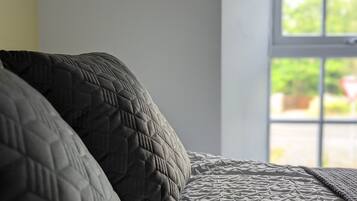 Una cama con un edredón gris texturizado y una almohada gris oscuro. Se observa una lámpara de pared con una bombilla visible. Se ve una ventana al fondo, mostrando follaje exterior.